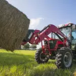 Mahindra 6075 cab utility tractor lifting hay bales in a grass field.