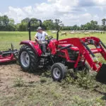 Mahindra 6075 utility tractor with bucket attachment driving in grass cattle field.