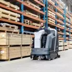 Operator driving Advance SC6000 rider scrubber through warehouse.