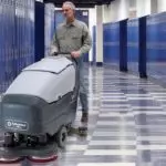 Operator walking behind Advance SC750 scrubber, cleaning school hallway.
