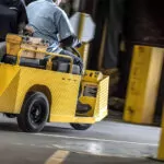 Cushman Minute Miser burden carrier driving out of a warehouse, showing passenger on rear seat.