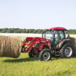 Mahindra 6075 tractor with cab transporting hay bales through open grass field.