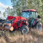 Mahindra 6075 cab utility tractor transporting logs with rake grapple attachment.