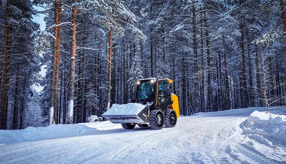 JCB 215 Skid Steer driving on snowy road with snow in bucket attachment.
