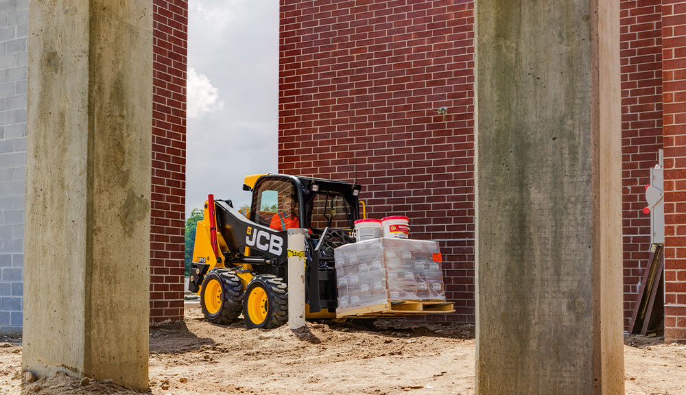 JCB 215 Skid Steer carrying pallet of materials at construction site.