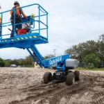 Workers driving Genie S-65 HF boom lift across rough dirt terrain.