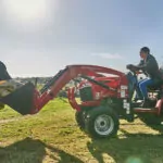 Mahindra MAX 26 XLT HST with bucket attachment lifting bags of animal food on a farm.