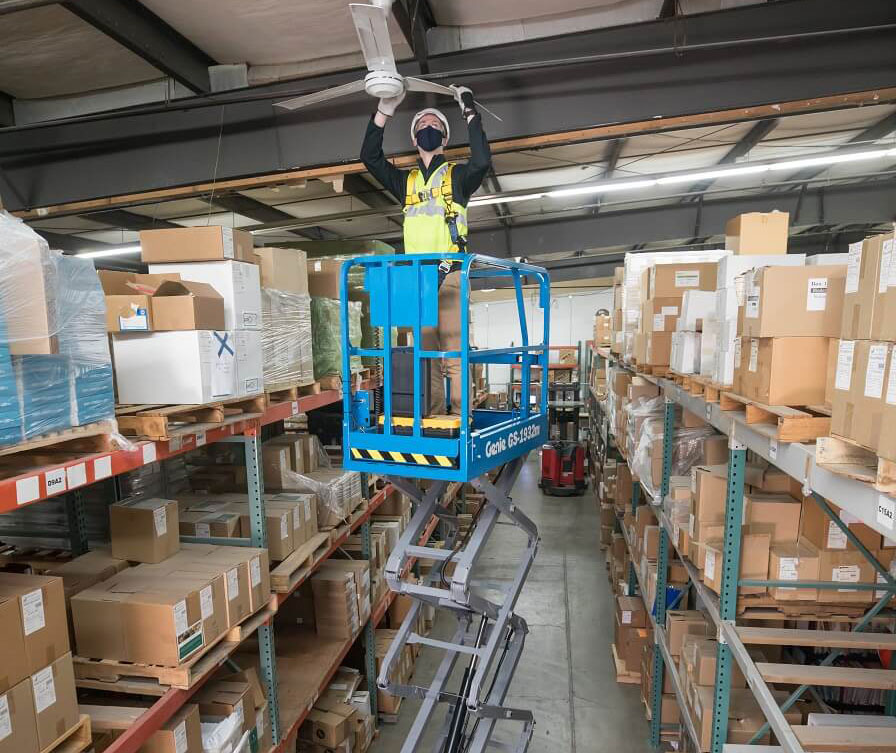 Genie GS 1932m scissor lift raising worker to a warehouse ceiling to perform cleaning maintenance.