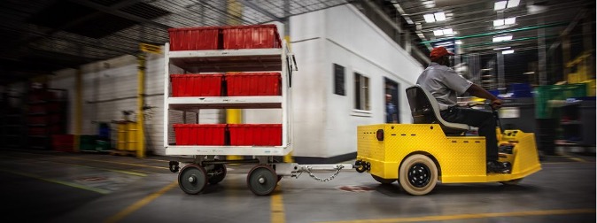 Tow tractor pulling cart with shelves and bins through warehouse.